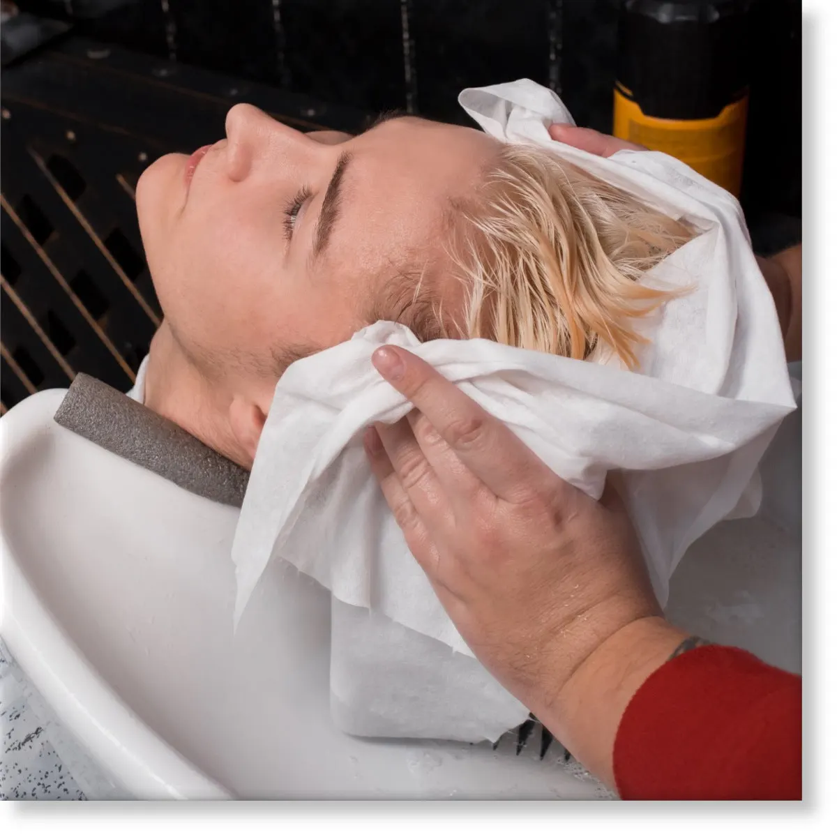 Hairdresser towel-drying a client's hair with a white disposable towel at the basin